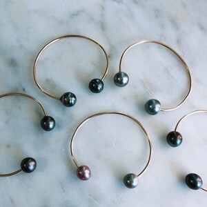 May include: Six gold-tone open cuff bracelets, each featuring a single dark pearl at each end. The pearls vary in color, including black, gray, and a reddish-brown hue. The bracelets are displayed on a white and gray marble surface.