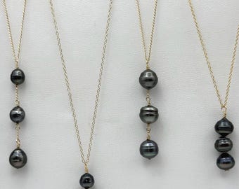 May include: Several gold chain necklaces with dark gray pearls of varying designs. Some necklaces feature a single pearl, while others have multiple pearls in a row. The pearls have a lustrous, smooth surface.