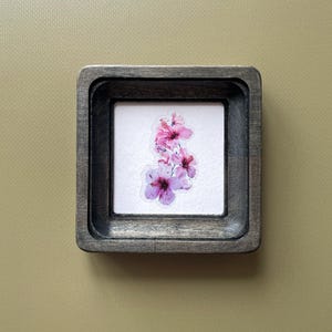 Pode incluir: Uma pequena obra de arte emoldurada em uma moldura de madeira quadrada. A moldura é de cor cinza escuro. Dentro da moldura há um fundo branco com um grupo de flores de hibisco rosa e roxo. As flores têm centros roxos escuros.