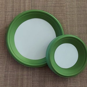 May include: Two green painted, round, wooden trays with white centers. The trays are different sizes, with the larger one partially overlapping the smaller one. The trays are on a brown, textured surface.