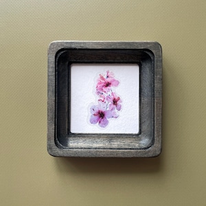 Puede incluir: Pequeña obra de arte enmarcada en un marco cuadrado de madera gris desgastada. La obra presenta un grupo de flores de hibisco rosas y moradas sobre un fondo blanco. El marco rodea el arreglo floral.