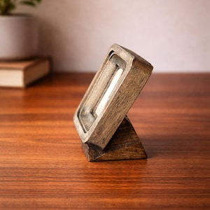 Puede incluir: Un peque&ntilde;o marco de fotos de madera en &aacute;ngulo con una abertura rectangular. El marco est&aacute; hecho de madera te&ntilde;ida oscura y tiene un dise&ntilde;o simple y minimalista. El marco est&aacute; apoyado en una peque&ntilde;a base triangular y est&aacute; sobre una superficie de madera.