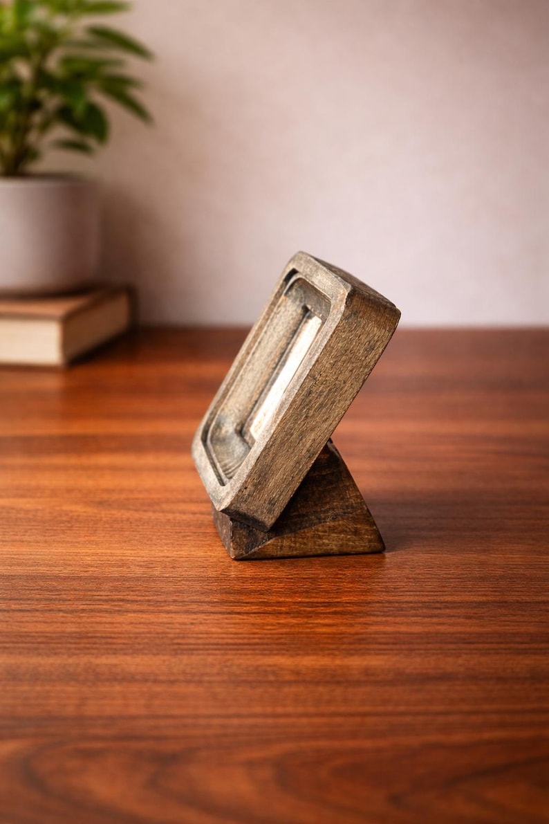 Puede incluir: Un peque&ntilde;o marco de fotos de madera en &aacute;ngulo con una abertura rectangular. El marco est&aacute; hecho de madera te&ntilde;ida oscura y tiene un dise&ntilde;o simple y minimalista. El marco est&aacute; apoyado en una peque&ntilde;a base triangular y est&aacute; sobre una superficie de madera.