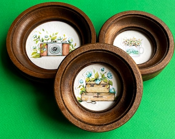 May include: Three wooden frames with decorative paper inserts. The frames are round and have a dark brown finish. The paper inserts feature watercolor illustrations of a camera, a bouquet of flowers, and a teacup.