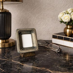 Puede incluir: Un peque&ntilde;o marco de fotos cuadrado de madera con un acabado dorado se encuentra sobre una superficie de m&aacute;rmol oscuro. El marco tiene un interior blanco y est&aacute; ligeramente inclinado. Un par de gafas descansa cerca, junto con un jarr&oacute;n dorado de rosas blancas.