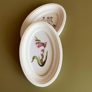 Puede incluir: Dos bandejas decorativas ovaladas blancas con diseños florales. Una bandeja presenta una flor rosa con hojas verdes, la otra flores rosas más pequeñas. Las bandejas están sobre un fondo verde apagado.