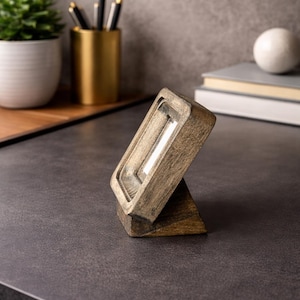 Puede incluir: Un marco de fotos de madera con un acabado envejecido, inclinado sobre un peque&ntilde;o soporte. El marco es rectangular con un &aacute;rea empotrada para una foto. El fondo incluye una planta, bol&iacute;grafos y libros.