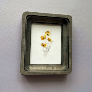 Puede incluir: Un pequeño marco rectangular de madera con un diseño en capas. En el interior, un fondo blanco muestra una delicada impresión botánica de flores amarillas con tallos verdes. El marco tiene un acabado gris envejecido.
