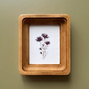 以下が含まれることがあります： 明るい茶色の仕上げの小さな正方形の木製フレーム。フレーム内には、濃い紫色の花のクラスターがある白い背景があります。フレームは、くすんだ緑色の背景に対して設定されています。