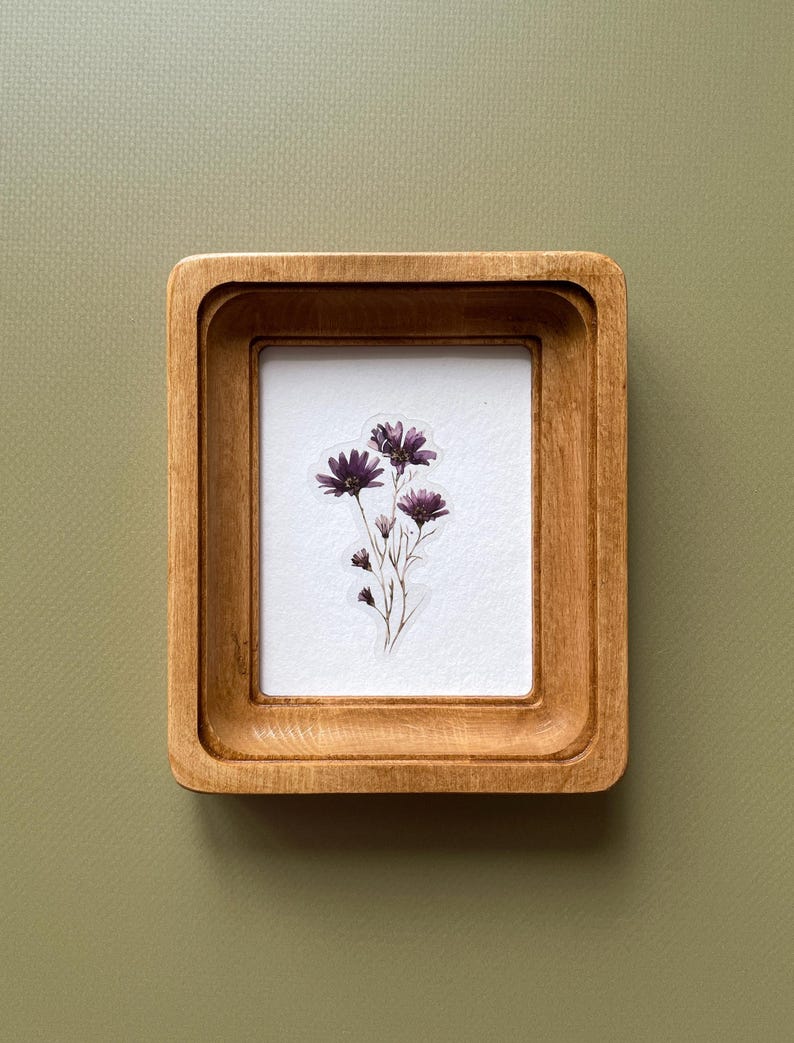 以下が含まれることがあります： 明るい茶色の仕上げの小さな正方形の木製フレーム。フレーム内には、濃い紫色の花のクラスターがある白い背景があります。フレームは、くすんだ緑色の背景に対して設定されています。