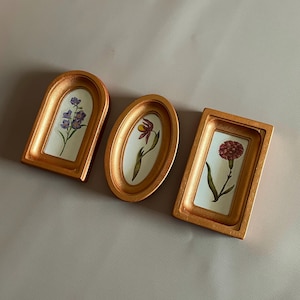 May include: Three decorative gold-colored frames, each with a different shape, showcasing botanical illustrations. The frames feature an arched, oval, and rectangular design, each holding a unique floral artwork with a white background.
