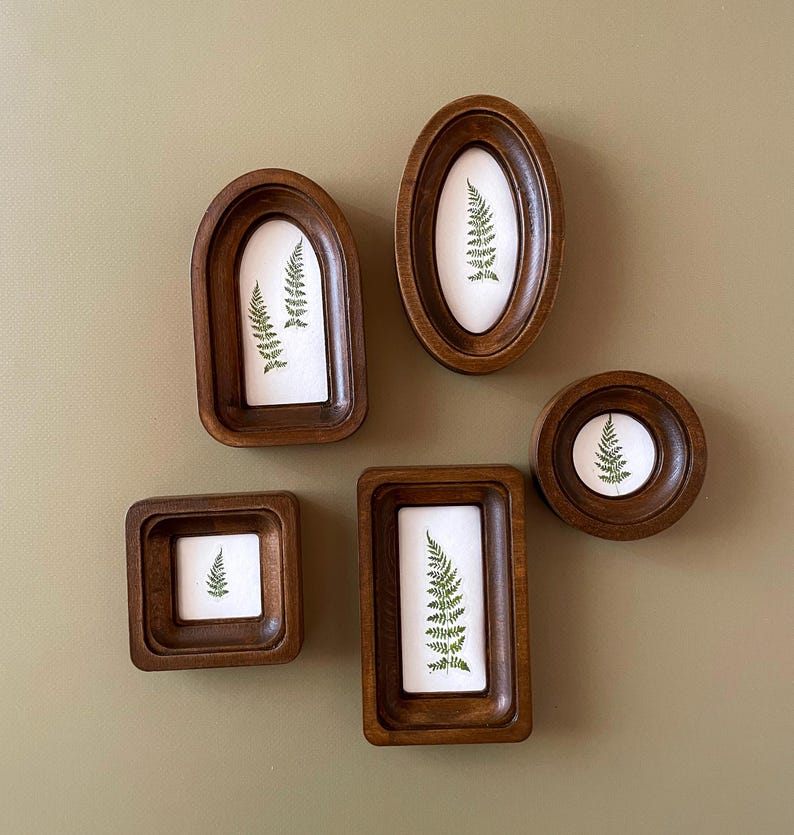 Puede incluir: Conjunto de cinco marcos de madera con estampados bot&aacute;nicos. Cada marco, de formas variadas (ovalado, redondo, cuadrado y rectangular), contiene una impresi&oacute;n de helecho verde sobre fondo blanco, creando una est&eacute;tica natural.
