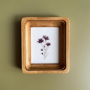以下が含まれることがあります： 明るい茶色の仕上げが施された、小さな長方形の木製フレーム。内側には、乾燥した紫色の花のクラスターが表示された白い紙があります。フレームはくすんだ緑色の背景に配置され、自然な美しさを生み出しています。