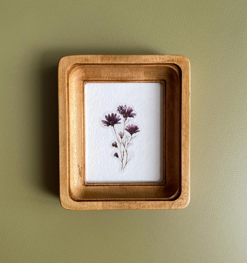 以下が含まれることがあります： 明るい茶色の仕上げが施された、小さな長方形の木製フレーム。内側には、乾燥した紫色の花のクラスターが表示された白い紙があります。フレームはくすんだ緑色の背景に配置され、自然な美しさを生み出しています。
