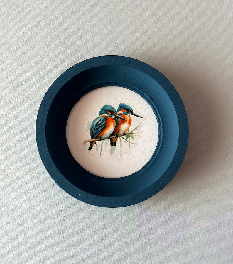May include: A blue round frame with a white background featuring a painting of two blue and orange birds perched on a branch.