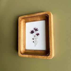 以下が含まれることがあります： 繊細な花の配置が施された、小さな木製の額装アート作品。 フレームは明るい茶色で、多層構造のデザインです。 アートワークは、白い背景に紫色の花のクラスターを展示し、自然でエレガントな美学を作り出しています。