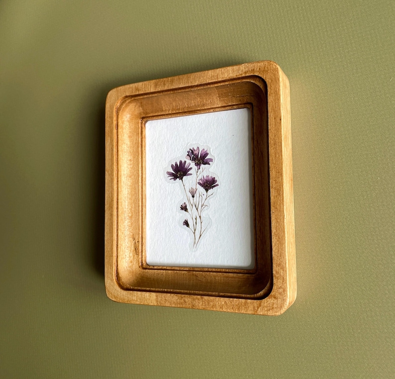 以下が含まれることがあります： 繊細な花の配置が施された、小さな木製の額装アート作品。 フレームは明るい茶色で、多層構造のデザインです。 アートワークは、白い背景に紫色の花のクラスターを展示し、自然でエレガントな美学を作り出しています。