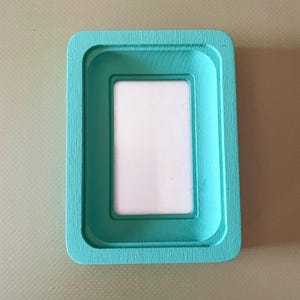 Può includere: Una cornice rettangolare in legno verniciato di turchese, con un bordo interno incassato e un inserto bianco. La cornice ha angoli arrotondati e una finitura liscia e verniciata. Progettata per contenere una piccola foto o opera d'arte.