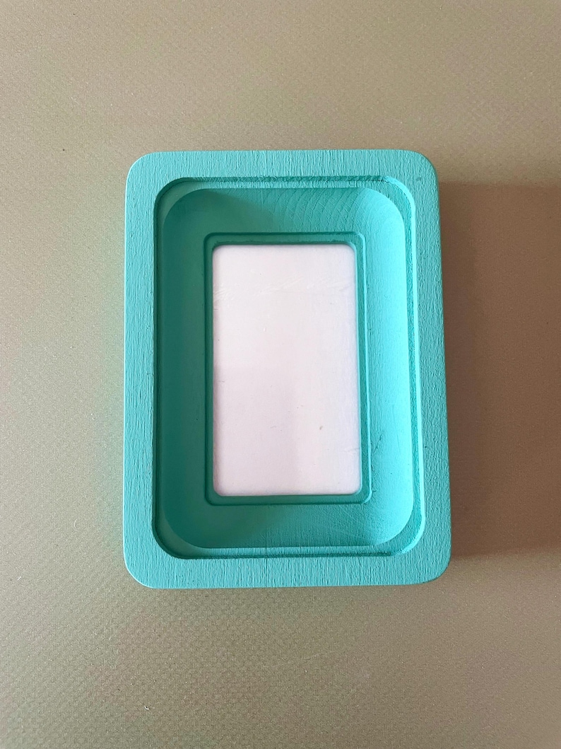 May include: A rectangular, turquoise-painted wooden picture frame with a recessed inner border and a white insert. The frame has rounded corners and a smooth, painted finish. The frame is designed to hold a small photograph or artwork.