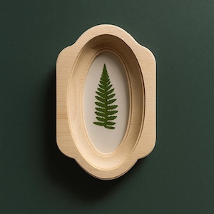 May include: A light-colored wooden frame with an oval opening, showcasing a green fern leaf against a cream-colored background. The frame has a decorative, scalloped edge. The background is a dark green.