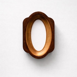 May include: A wooden oval picture frame with a decorative, scalloped edge. The frame is a warm brown colour with a lighter, golden-brown interior. The centre is a white oval, ready for a photo or artwork.