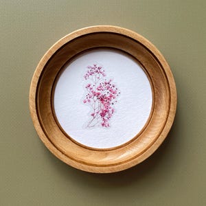 Op de afbeelding: Een ronde houten lijst met een lichtbruine afwerking, met een delicate compositie van roze gipskruidbloemen op een witte achtergrond. De lijst staat tegen een gedempte groene achtergrond.