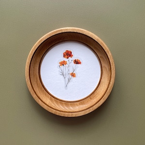 May include: A round wooden frame with a light brown finish. Inside the frame is a white background with a sticker of orange flowers. The frame is set against a muted green background.