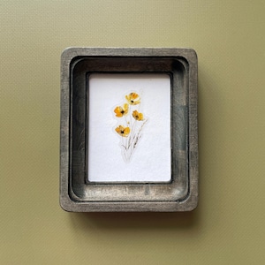 May include: A small, rectangular wooden frame with a gray finish. Inside, a white background displays a delicate botanical print of yellow flowers with dark centers and thin stems. The frame is set against a muted green backdrop.