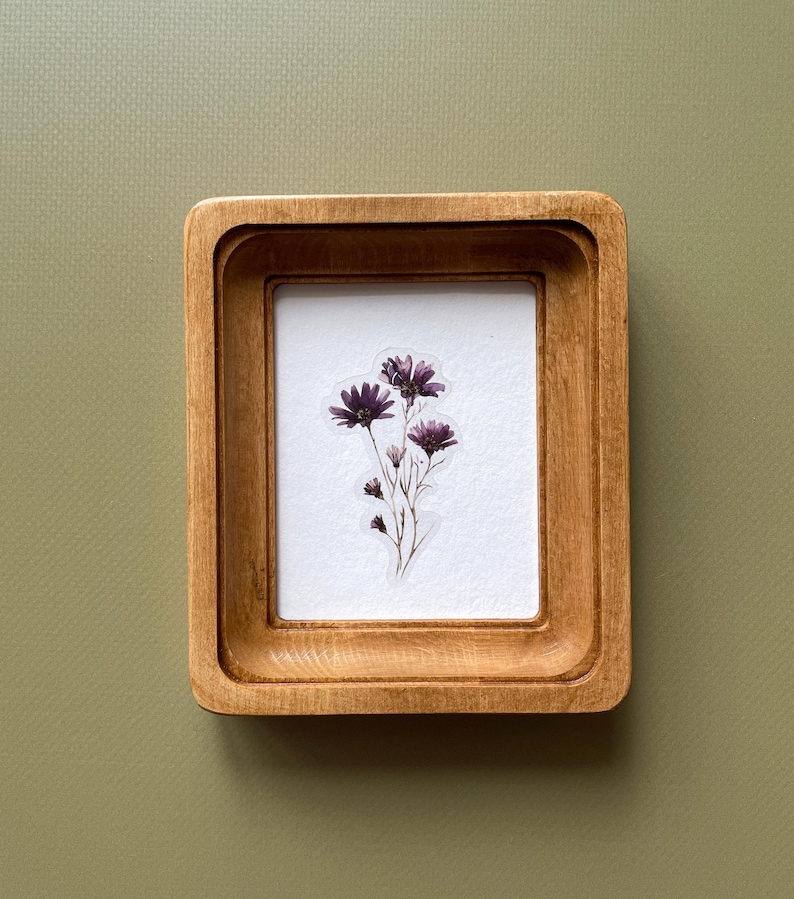 以下が含まれることがあります： 明るい茶色の仕上げが施された、小さな長方形の木製フレーム。フレーム内には、紫色の花の繊細なアレンジメントが施された白い背景があります。フレームは、くすんだ緑色の背景に配置されています。