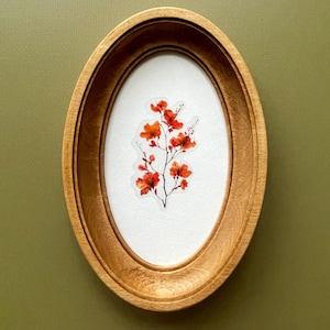 Könnte beinhalten: Ein ovaler Holzrahmen mit einem botanischen Druck von roten Blumen auf weißem Hintergrund. Der hellbraune Rahmen ist an einer grünen Wand befestigt. Das Kunstwerk ist eine zarte und naturalistische Darstellung floraler Elemente, geeignet für die Dekoration.