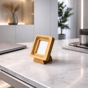 May include: A small, square wooden picture frame with a light-colored finish. The frame is angled and stands on a small base. The frame is empty, with a white interior. The frame is on a marble-like surface.