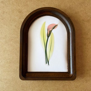May include: A framed print of a single pink calla lily with green leaves on a white background.