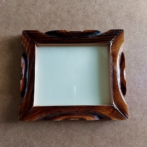 May include: A small, rectangular wooden picture frame with a dark brown finish. The frame has a carved, ornate design on the corners and sides, and a light-colored interior. The frame is empty.