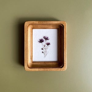 以下が含まれることがあります： 小さな長方形の金色の木製フレームと白いマット。 フレームには、紫色の花の繊細な植物プリントが収められています。 背景は無地のオリーブグリーンです。