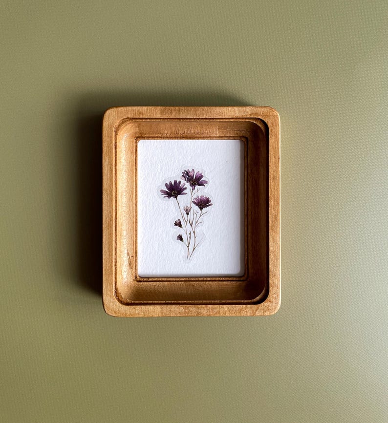 以下が含まれることがあります： 小さな長方形の金色の木製フレームと白いマット。 フレームには、紫色の花の繊細な植物プリントが収められています。 背景は無地のオリーブグリーンです。