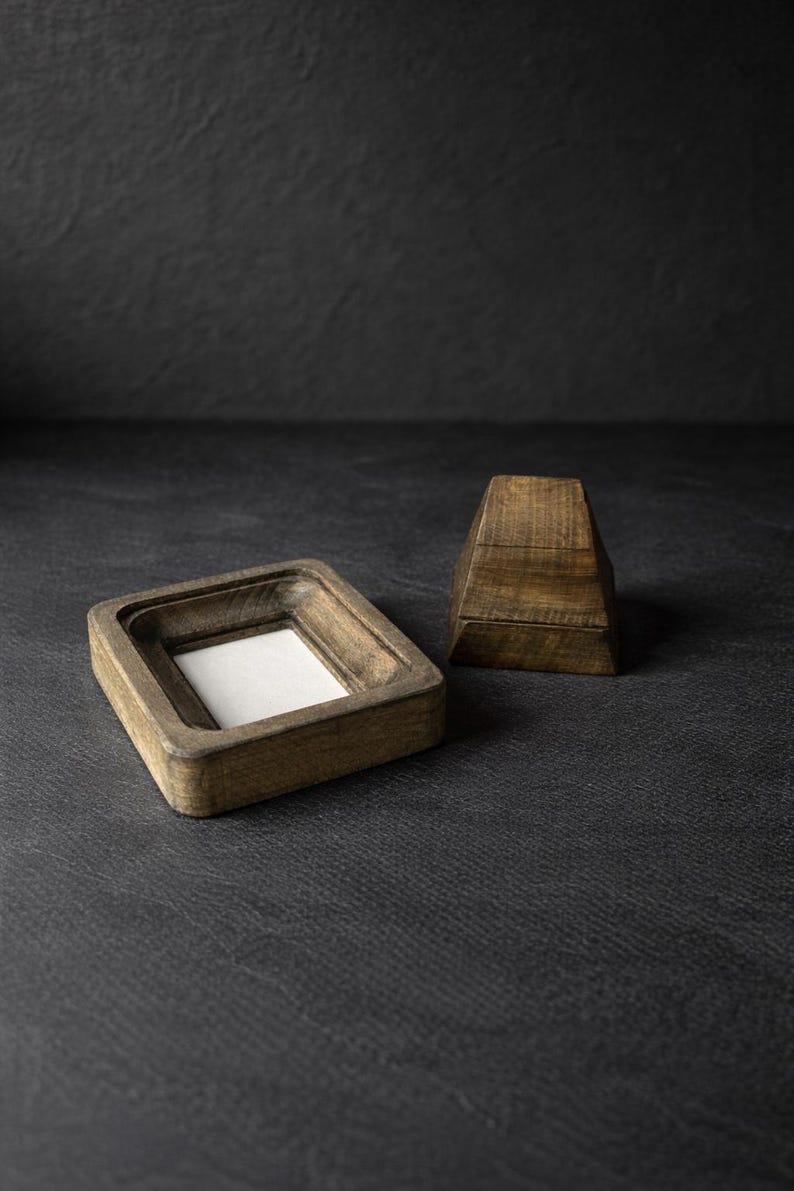 Puede incluir: Un marco de fotos de madera con un inserto blanco y un peque&ntilde;o objeto geom&eacute;trico de madera. El marco es cuadrado con un &aacute;rea empotrada para la foto. Los objetos son de madera de color marr&oacute;n claro y est&aacute;n sobre una superficie oscura.