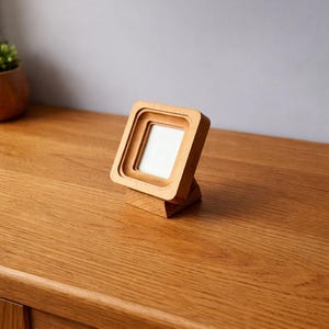 May include: A small, square wooden picture frame with a layered design. The frame is made of light-colored wood and stands upright on a matching base. The frame is empty, with a white interior. The frame is on a wooden surface.