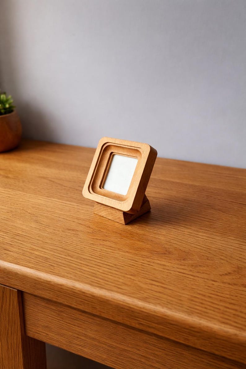 May include: A small, square wooden picture frame with a layered design. The frame is made of light-colored wood and stands upright on a matching base. The frame is empty, with a white interior. The frame is on a wooden surface.