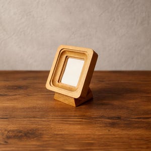 May include: A small, square wooden picture frame with a light-colored finish. The frame has a layered design and is set on a wooden base. The background is a neutral gray wall and a dark brown wooden surface.