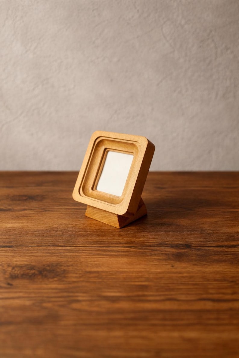 May include: A small, square wooden picture frame with a light-colored finish. The frame has a layered design and is set on a wooden base. The background is a neutral gray wall and a dark brown wooden surface.