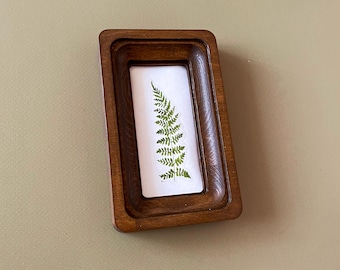 Puede incluir: Un pequeño marco rectangular de madera con un acabado marrón oscuro. En el interior, un fondo blanco muestra un helecho verde prensado. El marco tiene un borde biselado, lo que añade un toque de encanto vintage. La estética general es natural y botánica.
