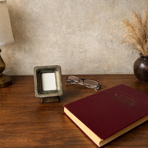 Puede incluir: Un peque&ntilde;o marco de fotos cuadrado de madera con un inserto blanco est&aacute; sobre una superficie de madera. Un par de gafas y un libro granate tambi&eacute;n est&aacute;n sobre la superficie. Una l&aacute;mpara y un jarr&oacute;n con plantas secas est&aacute;n en el fondo.