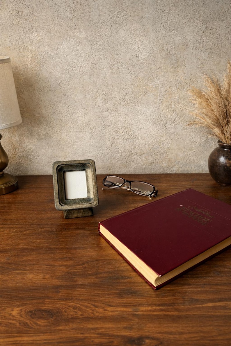 Puede incluir: Un peque&ntilde;o marco de fotos cuadrado de madera con un inserto blanco est&aacute; sobre una superficie de madera. Un par de gafas y un libro granate tambi&eacute;n est&aacute;n sobre la superficie. Una l&aacute;mpara y un jarr&oacute;n con plantas secas est&aacute;n en el fondo.