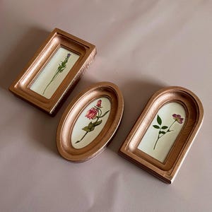 Puede incluir: Tres marcos de flores prensadas de color cobre. Los marcos son rectangulares, ovalados y arqueados. Las flores incluyen una ramita de lavanda, una rosa y una flor morada. Los marcos están sobre una superficie rosa.