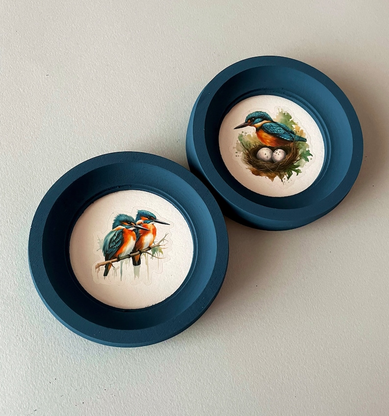 May include: Two dark blue round picture frames with white mats. One frame features a painting of two kingfishers perched on a branch. The other frame features a painting of a kingfisher sitting on a nest with eggs.