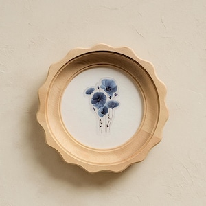 May include: A round, natural wood frame with a scalloped edge. Inside the frame is a white background with a cluster of blue floral artwork. The artwork features delicate, translucent flowers.
