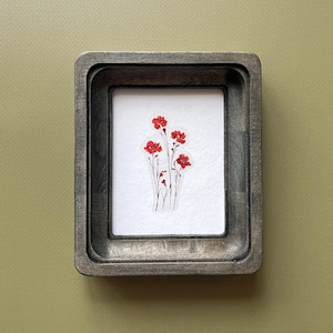 May include: A framed artwork featuring a cluster of red flowers on a white background. The artwork is encased in a dark gray, rectangular wooden frame. The background is a muted olive green.