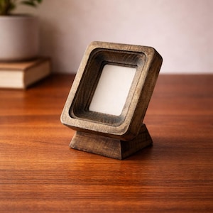 Puede incluir: Un pequeño marco de fotos de madera en ángulo con una abertura cuadrada. El marco está hecho de madera teñida oscura y se asienta sobre una pequeña base. El marco está vacío, con un interior blanco. El marco se muestra sobre una superficie de madera.