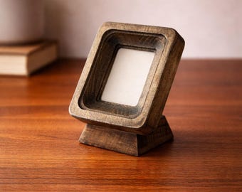 May include: A small, angled wooden picture frame with a square opening. The frame is made of dark-stained wood and sits on a small base. The frame is empty, with a white interior. The frame is displayed on a wooden surface.