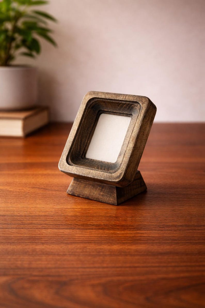 Puede incluir: Un peque&ntilde;o marco de fotos de madera en &aacute;ngulo con una abertura cuadrada. El marco est&aacute; hecho de madera te&ntilde;ida oscura y se asienta sobre una peque&ntilde;a base. El marco est&aacute; vac&iacute;o, con un interior blanco. El marco se muestra sobre una superficie de madera.